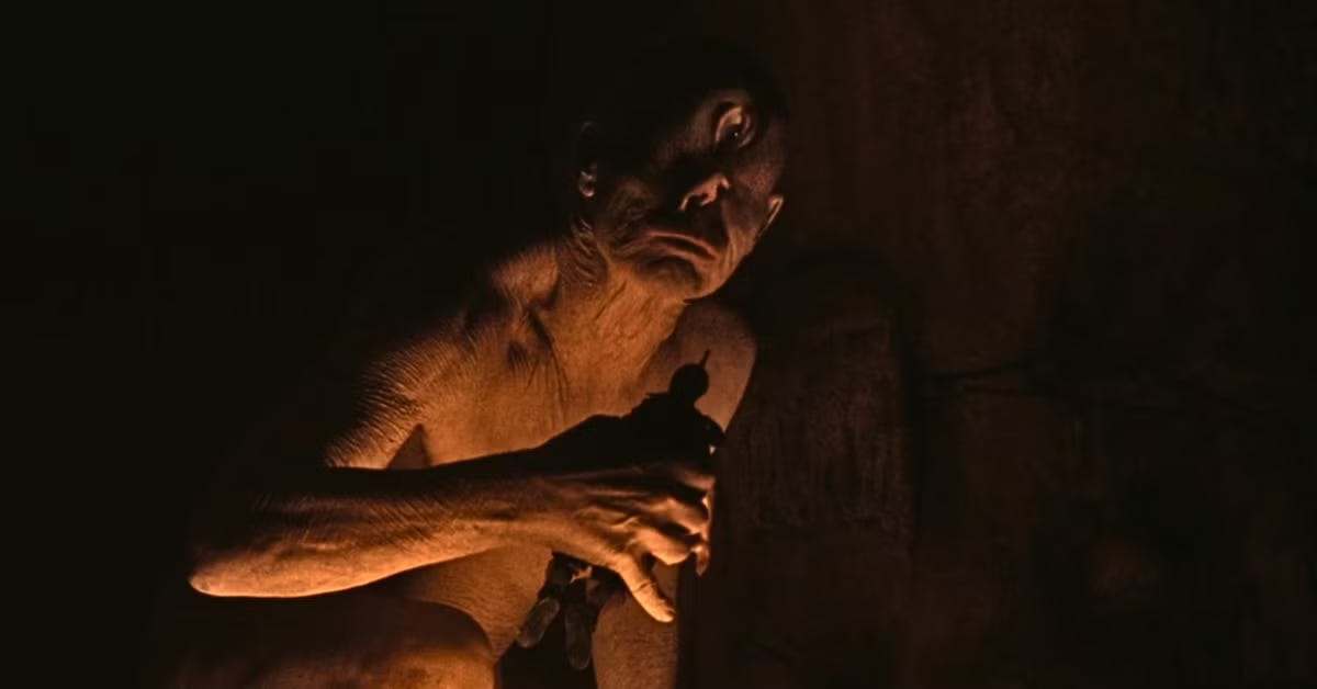 Movie still from The Odyssey. A one-eyed cyclops monster sits in a dark cave lit by a fire.