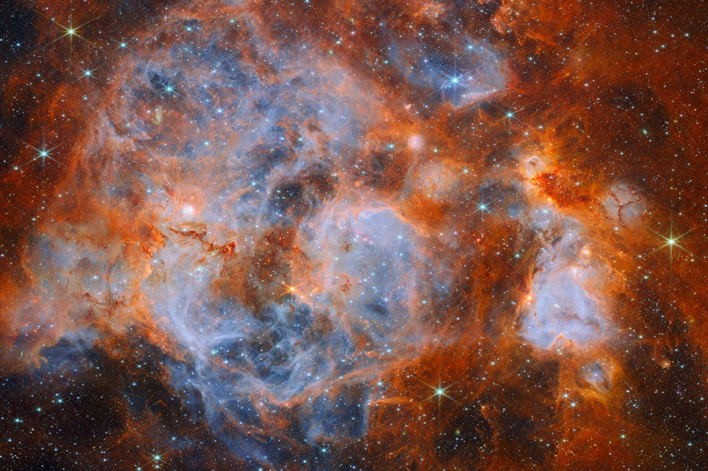 A nebula made of clouds of gas and dust with stars forming within. In the center and out to the left it is filled with thin, wispy blue gas that forms delicate layers. Around this are thicker orange clouds with dark ridges. Thin red filaments of gas cross the center of the nebula. Glowing bubbles in the gas contain dense, very bright clusters of stars. In the edges, distant stars are visible as bright points on a dark background. A nebula made of clouds of gas and dust with stars forming within. In the center and out to the left it is filled with thin, wispy blue gas that forms delicate layers. Around this are thicker orange clouds with dark ridges. Thin red filaments of gas cross the center of the nebula. Glowing bubbles in the gas contain dense, very bright clusters of stars. In the edges, distant stars are visible as bright points on a dark background.