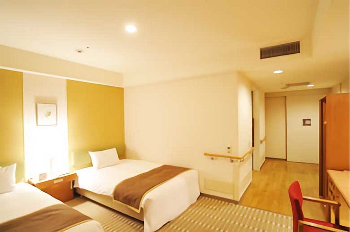 A collage of four photos of Hotel the Lutheran in Osaka. The top left shows the modern exterior entrance lit warmly at night. The top right shows a bright, minimalist guest room with a double bed, wooden floors, and simple furnishings. The bottom left highlights a wide hallway with handrails along the wall, suitable for accessibility needs. The bottom right shows a spacious accessible bathroom with grab bars, a roll-in shower with a curtain, and a toilet designed for ease of use.