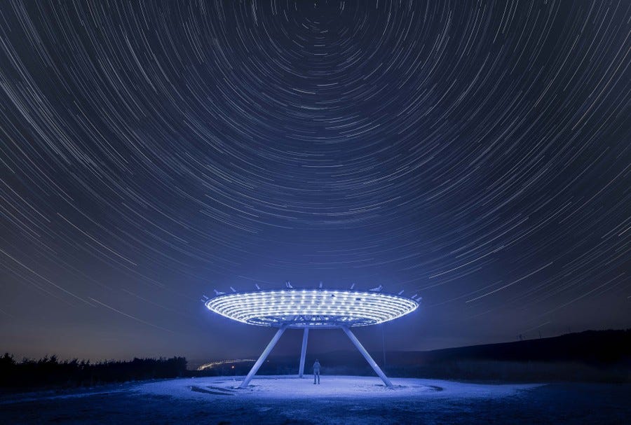 A long-exposure photo shows star trails forming circles above an illuminated sculpture on a hill. A long-exposure photo shows star trails forming circles above an illuminated sculpture on a hill.