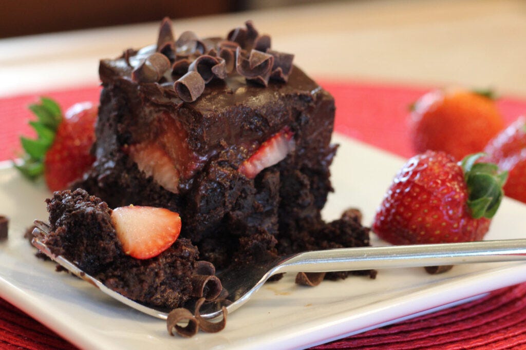 Stylized photo of chocolate ganache and strawberry brownies featuring a brownie on a plate, fork filled with brownie bite and strawberry, fresh strawberries surround the plate.