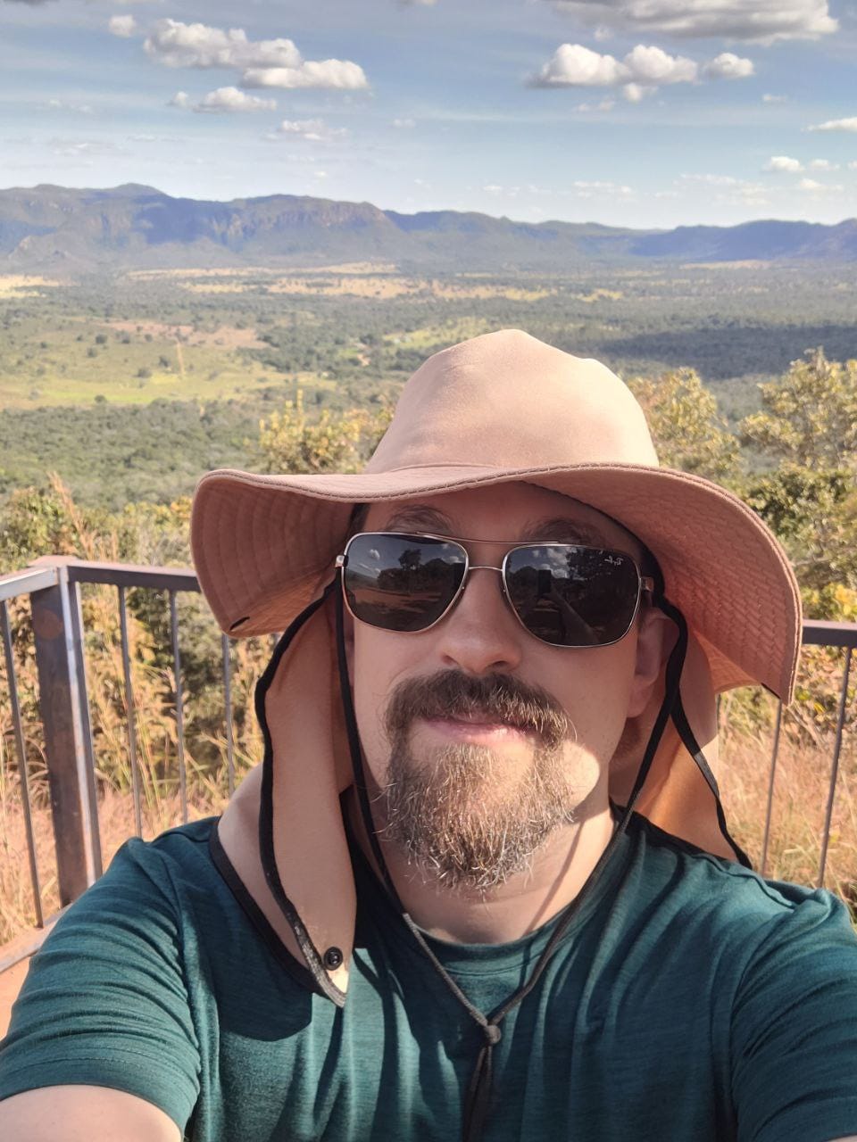 Eu, homem branco de cavanhaque, óculos escuros, camiseta verde escuro, um chapéu daqueles que cobrem o pescoço, tirando uma selfie numa grande com um grande vale do Cerrado ao fundo.