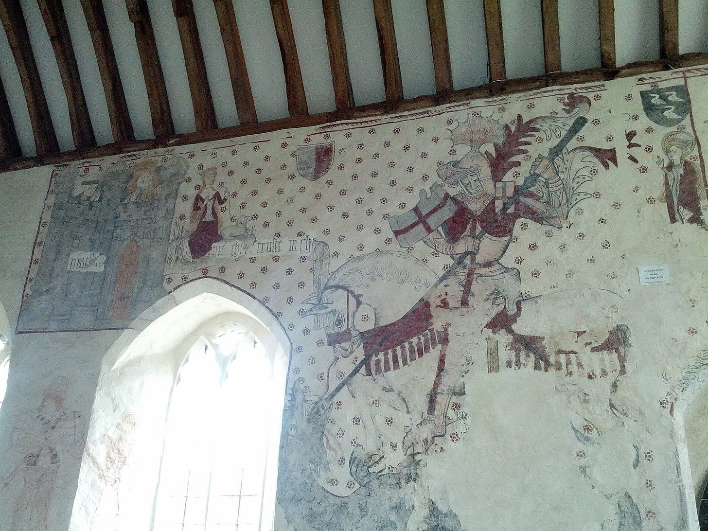 A church wall under wooden rafters, painted with a huge knight in armour on a white horse, red cross banner and lance, a castle with two crowned figures at the top left and a woman with a lamb, on a background scattered with small red flowers. A church wall under wooden rafters, painted with a huge knight in armour on a white horse, red cross banner and lance, a castle with two crowned figures at the top left and a woman with a lamb, on a background scattered with small red flowers.