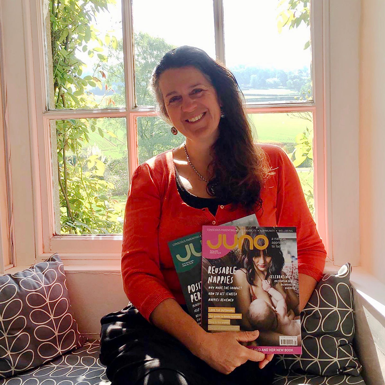 Editor Saffia Farr wearing a bright coral top holding copies of JUNO, against a sunny window with greenery beyond Editor Saffia Farr wearing a bright coral top holding copies of JUNO, against a sunny window with greenery beyond