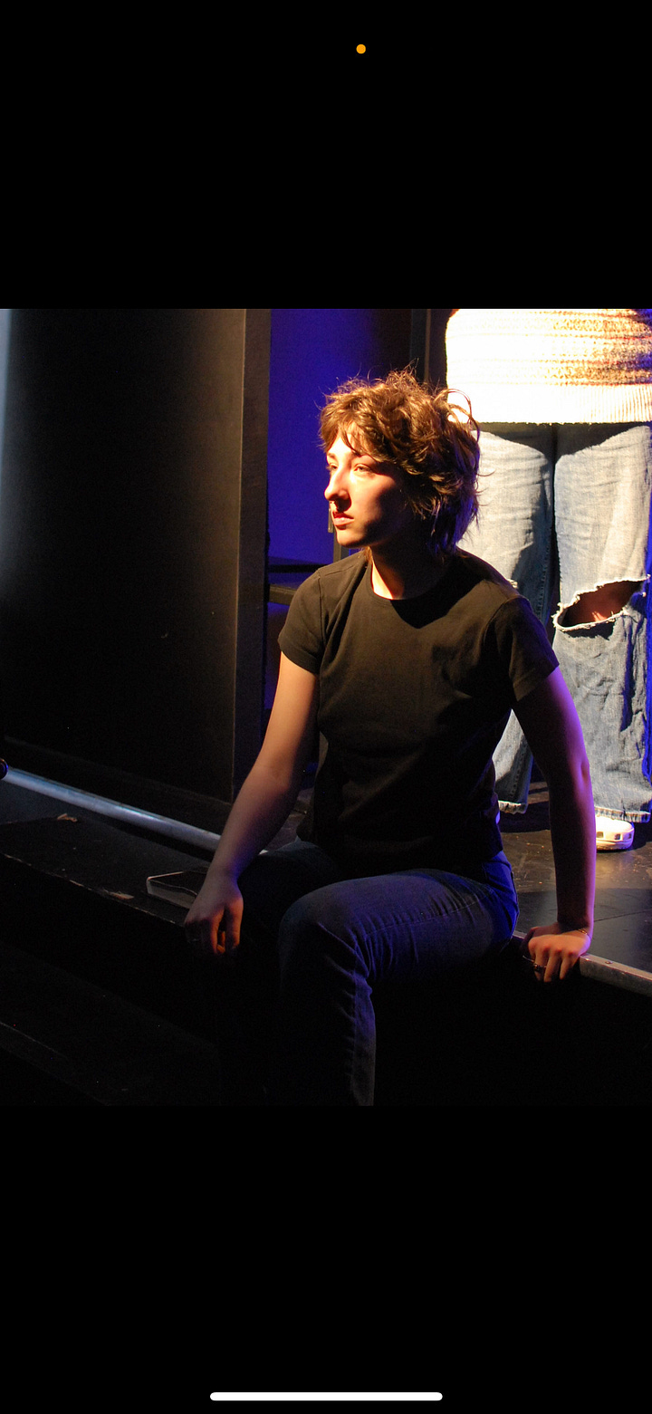 A person with short, tousled brown hair sits on the edge of a stage under dramatic lighting. They wear a plain black t-shirt and jeans, gazing pensively toward a light source. The scene is dimly lit with a deep blue and purple backdrop, and another person stands behind them, visible only from the waist down, wearing jeans with a large knee tear. The atmosphere suggests a theatrical or emotional performance moment. On the rightA black and white photograph of a person seated on a chair at the edge of a stage, focused intently on writing in a notebook balanced on their lap. They wear a dark t-shirt, jeans, and Adidas sneakers. Soft light falls on their face and hands, creating a contemplative atmosphere. In the blurred background, another figure sits quietly in shadow, suggesting a behind-the-scenes or rehearsal setting in a theater.