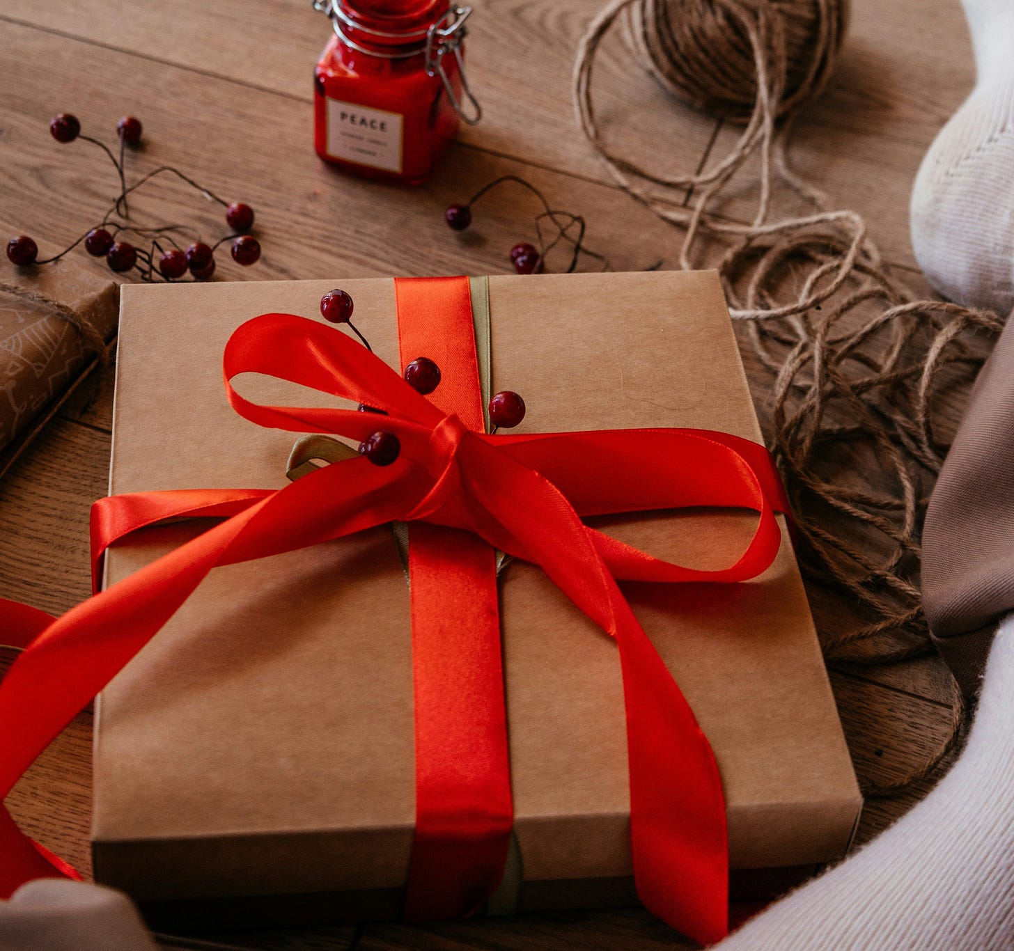 Photo shows a gift package wrapped in brown paper and red fabric ribbon, accented with some faux red berries.  In the background is a wood table with various wrapping supplies scattered around.