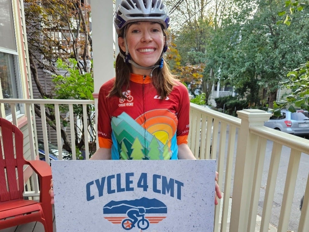A young woman holds a sign that says CYCLE 4 CMT while wearing a bike helmet. A young woman holds a sign that says CYCLE 4 CMT while wearing a bike helmet.