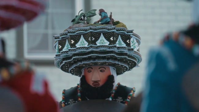 Un Suisse ave un énorme couvre-chef et le visage recouvert d'un masque figé.