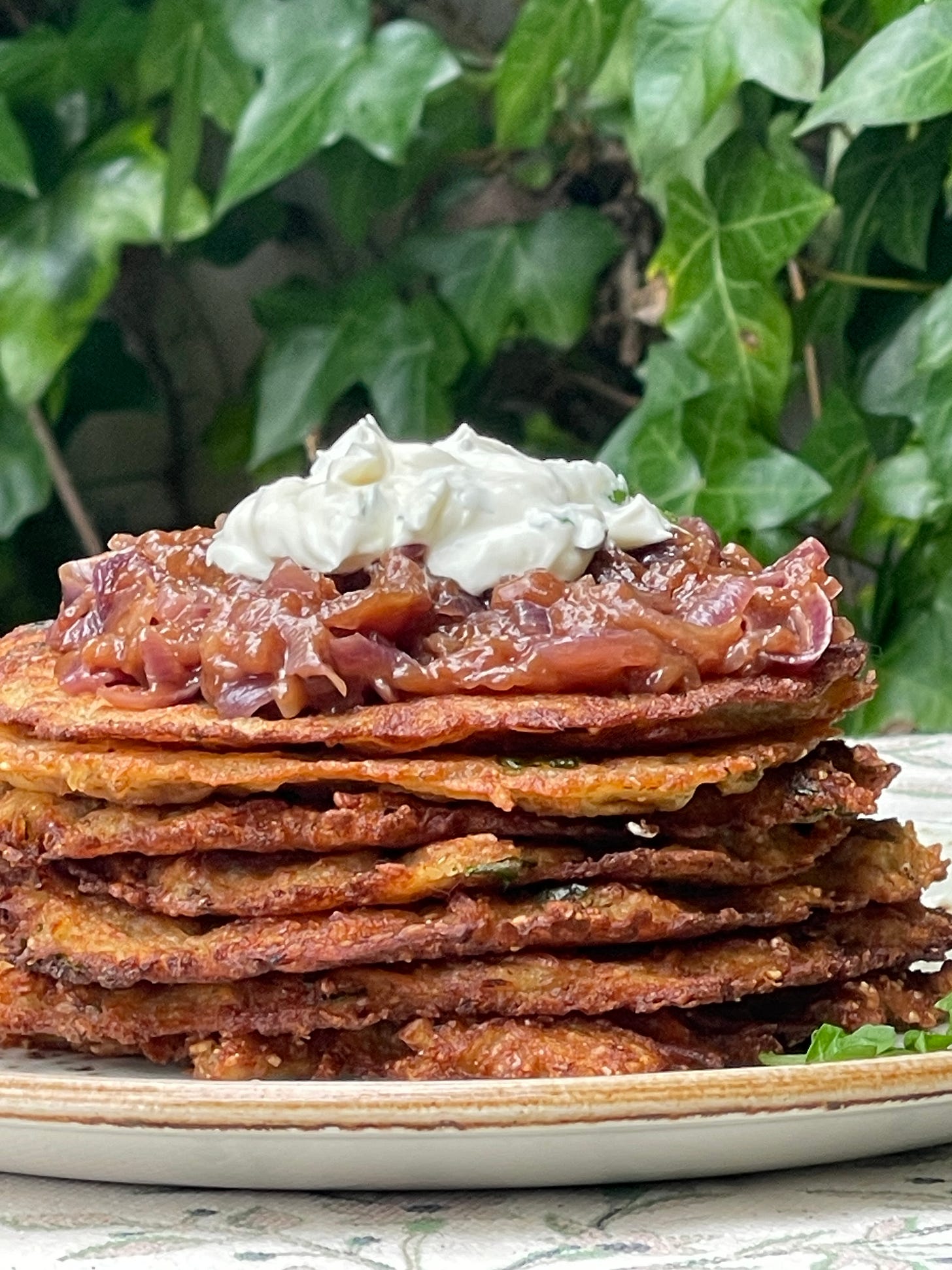 A tall stack of crisp bramboráky — Czech garlic potato pancakes — topped with beer-caramelized red onions and herbed cream, served on a ceramic plate against a backdrop of trailing ivy. A tall stack of crisp bramboráky — Czech garlic potato pancakes — topped with beer-caramelized red onions and herbed cream, served on a ceramic plate against a backdrop of trailing ivy.