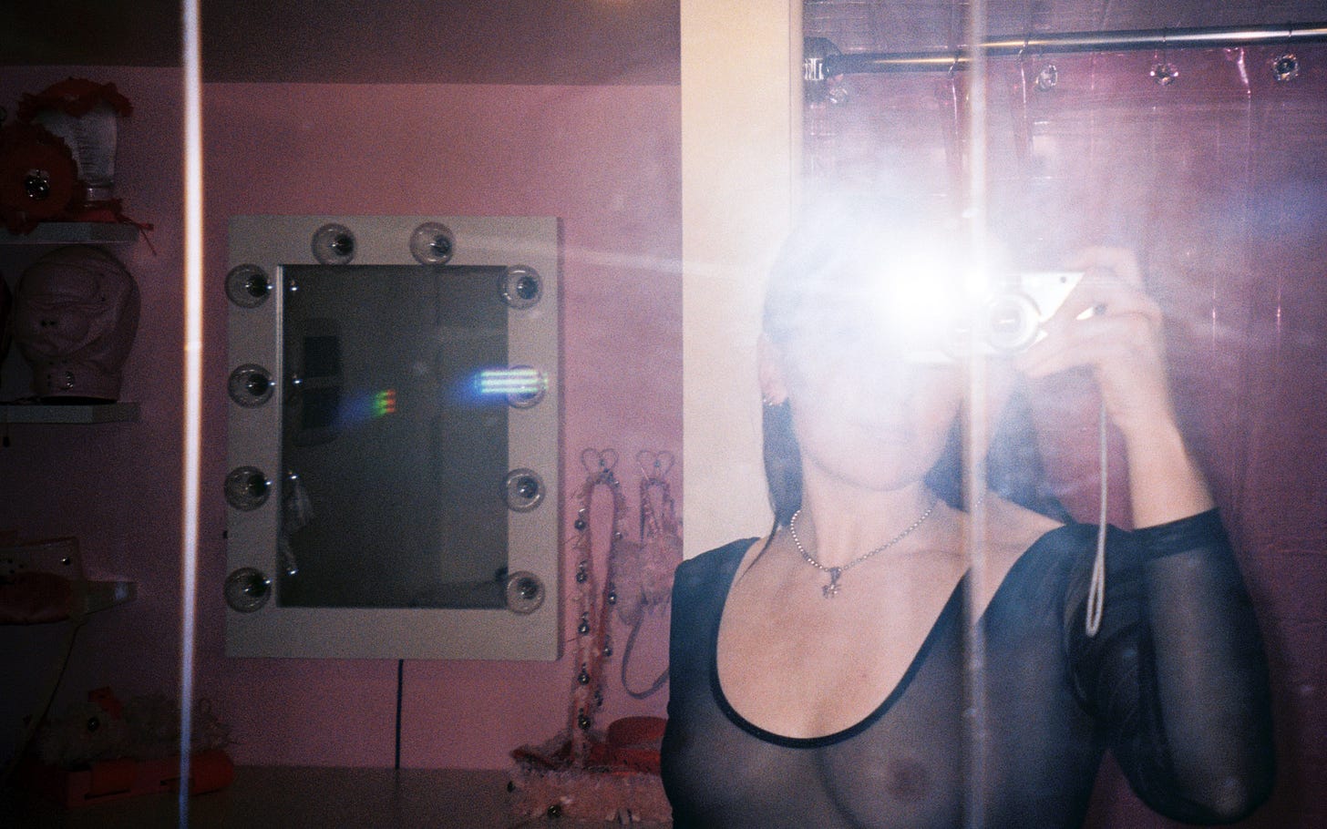 A film mirror selfie of Evyn Silver, a white femme with long dark hair, wearing a black mesh long sleeve top and a silver necklace with a spider pendant. The flash from the camera obscures her face. In the background is a pink bathroom with a pink plastic shower curtain and a vanity with lights around the mirror. Above the vanity are shelves displaying pink hoods and ruffled accessories.