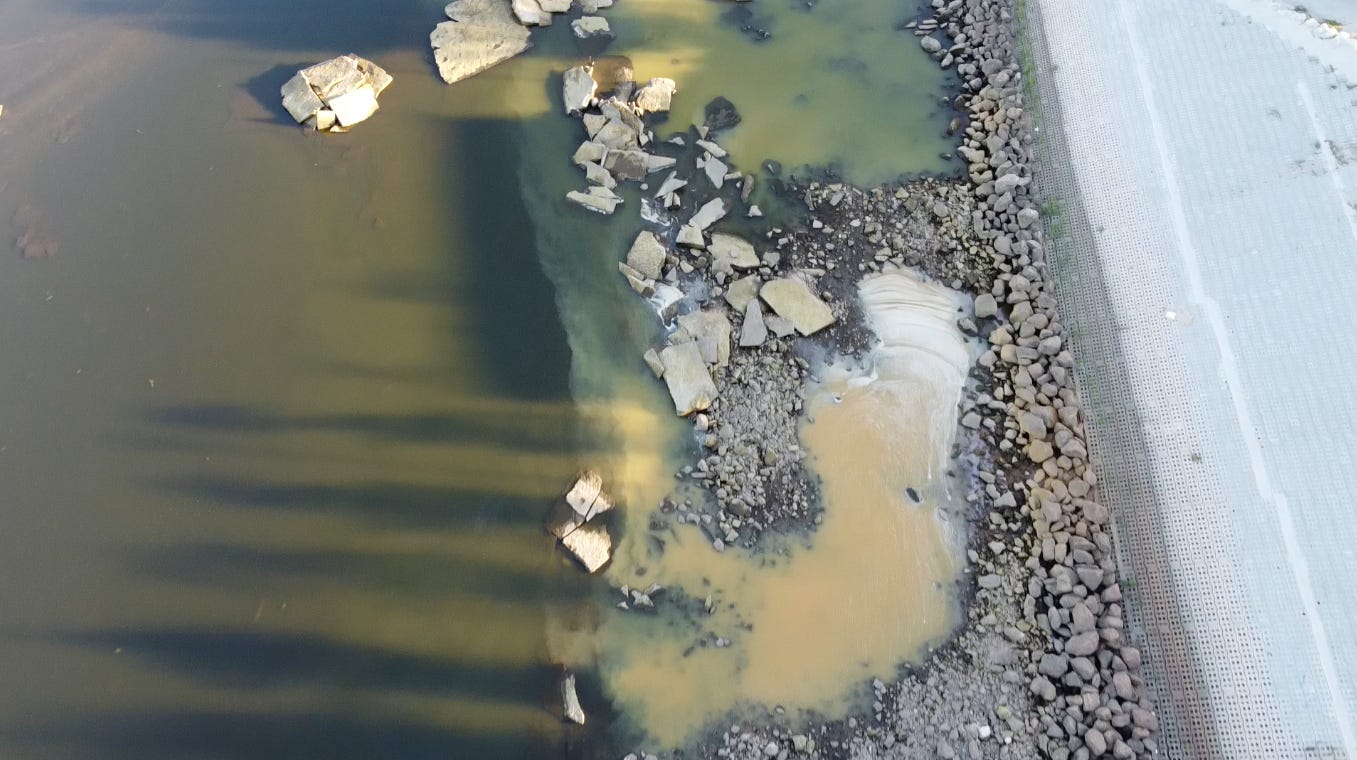 Photographed from an overhead drone, long shadows of trees cross plumes of green and orange water billowing into the dark green channel of the Arkansas River from a rocky shoreline and concrete embankment.