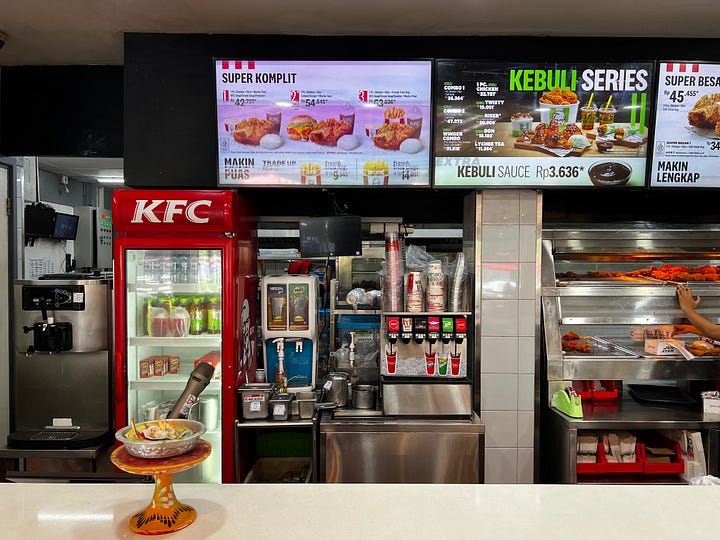  The interior of a KFC, customers at fixed stools, large letters on the wall reading HARVEST and below it NO WASTE NO HUNGER. KFC menu boards advertising rice meal sets, a Kebuli Series, and lychee tea above the service counter. 