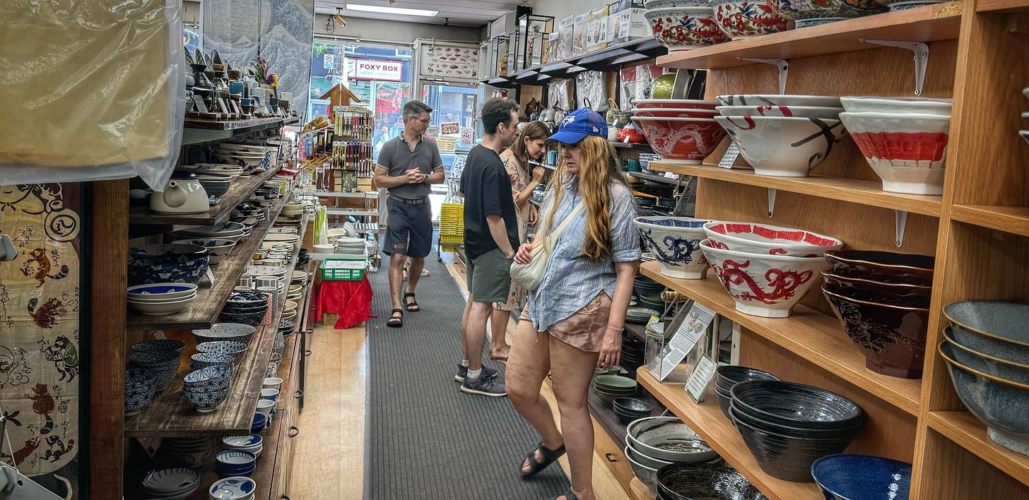 The housewares aisle inside Sanko. On the shelves are a variety of patterned bowls and dishes. All of the shoppers are White.