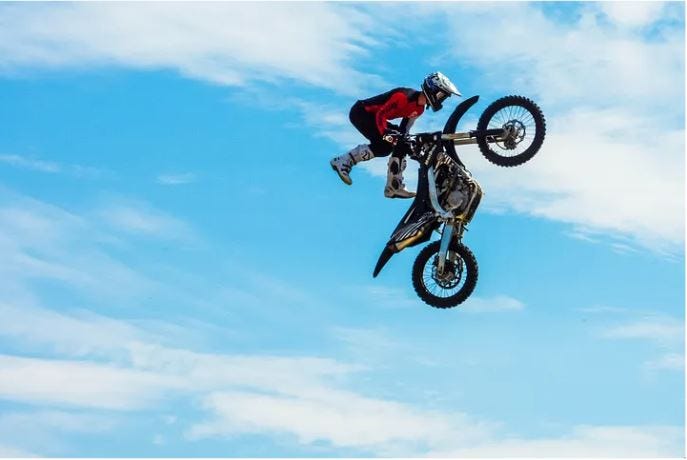 Motorbike rider in red with white helmet and bootsdoing a trick in the air against blue sky with white clouds Motorbike rider in red with white helmet and bootsdoing a trick in the air against blue sky with white clouds