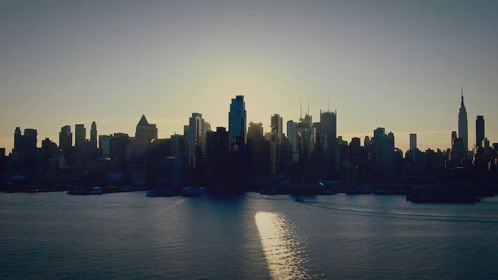 A wide shot of a city skyline—likely New York—seen across the water at dusk, with the sun setting behind the buildings and a streak of reflected light stretching across the surface.