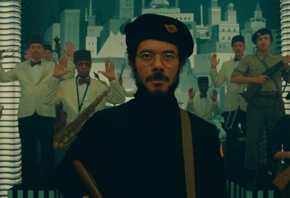 Actor Richard Ayoade as his character, a communist revolutionary, in the Phoenician Scheme. He is dressed in all black, carrying a gun, and wearing a beret. Behind him people have their hands up.