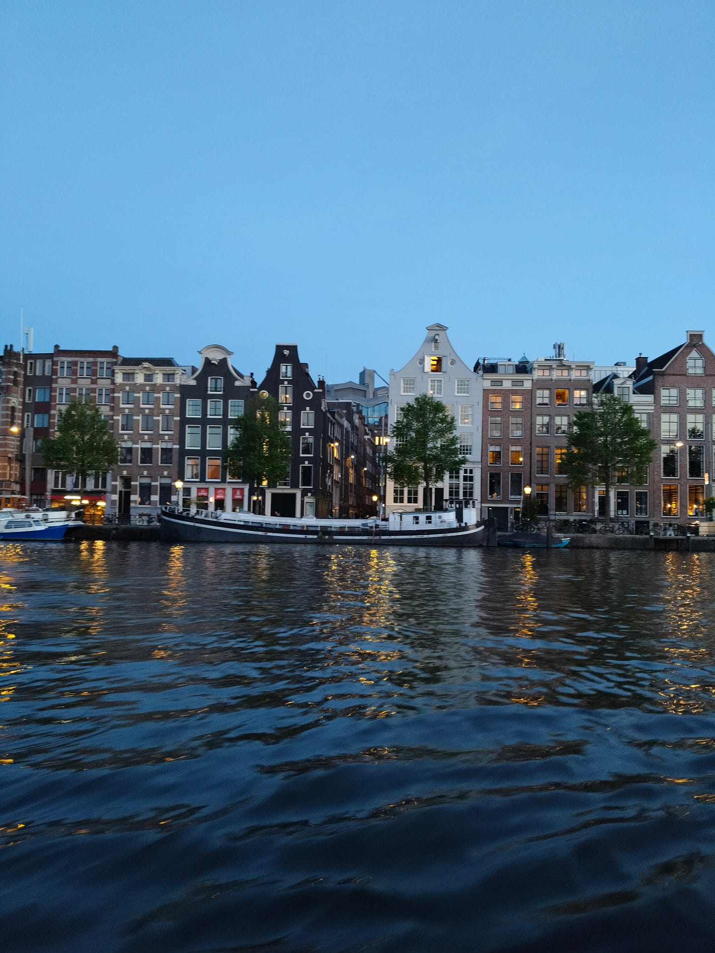 A view from the water: tall, skinny houses kiss the edge of the canal at dusk. A darkening clear blue sky hangs above. Old trailer boats are moored. 