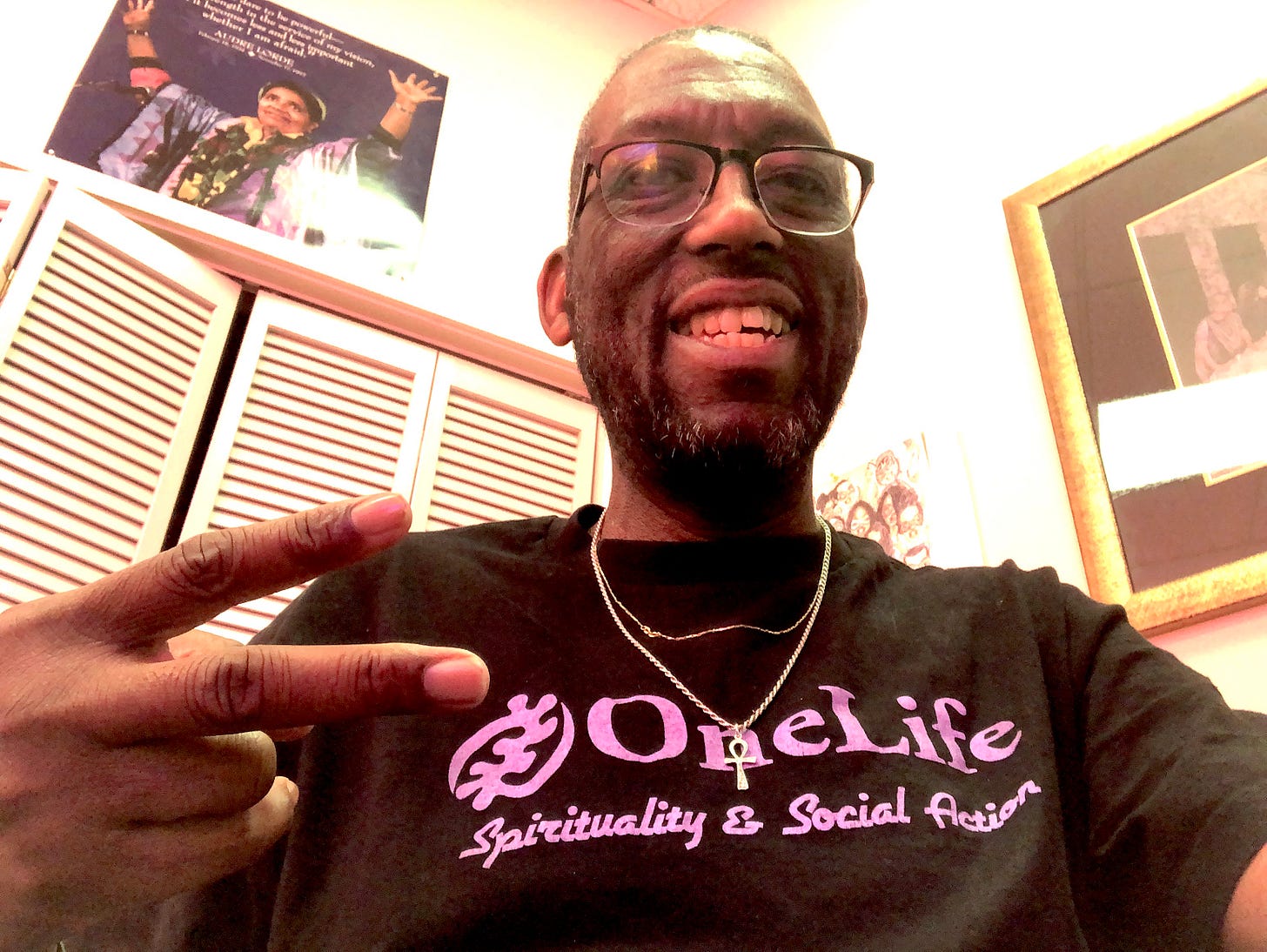 Kamal in his office wearing a black OneLife Institute T-shirt with lavender printed slogan "Spirituality & Social Action." He is smiling and flashing a peace sign, behind him is a poster of Audre Lorde.
