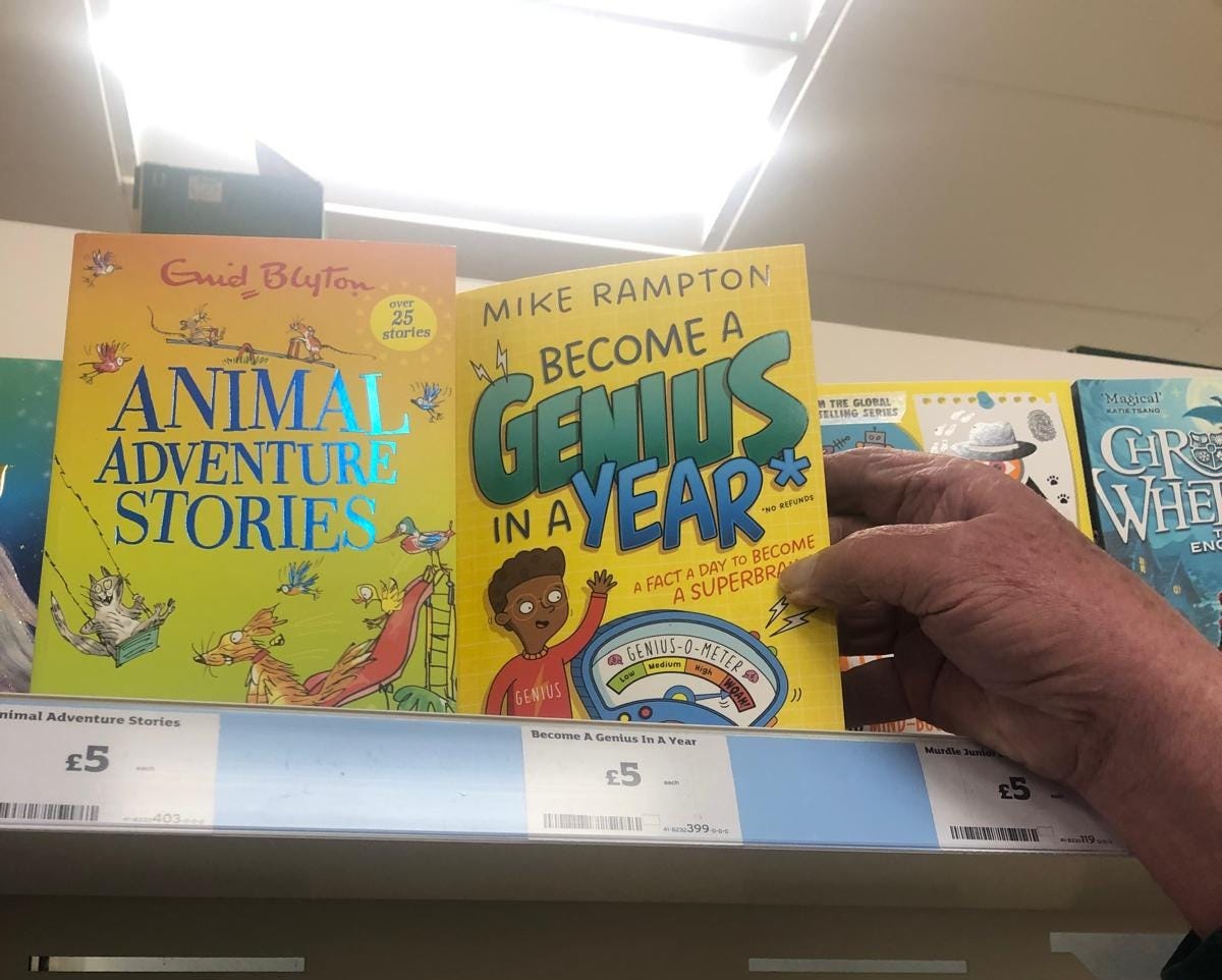 A hand (my dad's) reaching for Become A Genius In A Year on a shelf in Sainsbury's in Coleraine. It is shelved next to an Enid Blyton compilation.