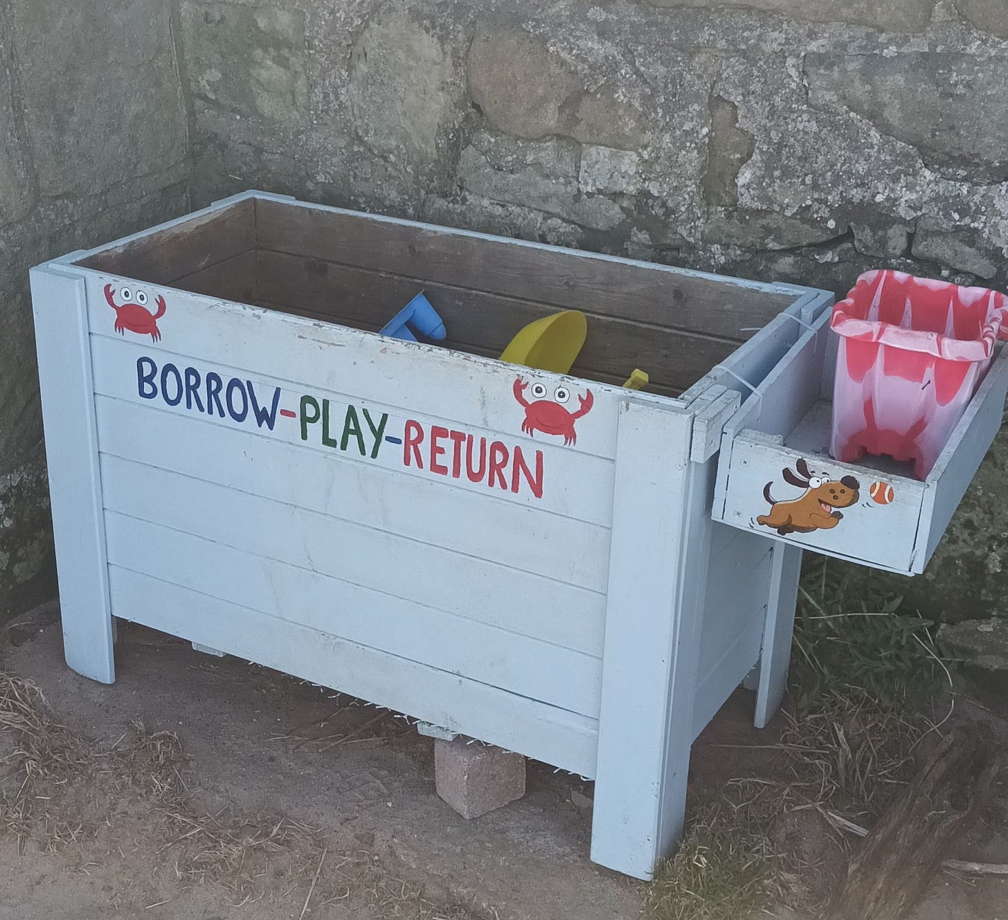 'Toy Library' on the beach, a wooden box painted white with the words 'borrow-play-return' painted on and beach toys inside