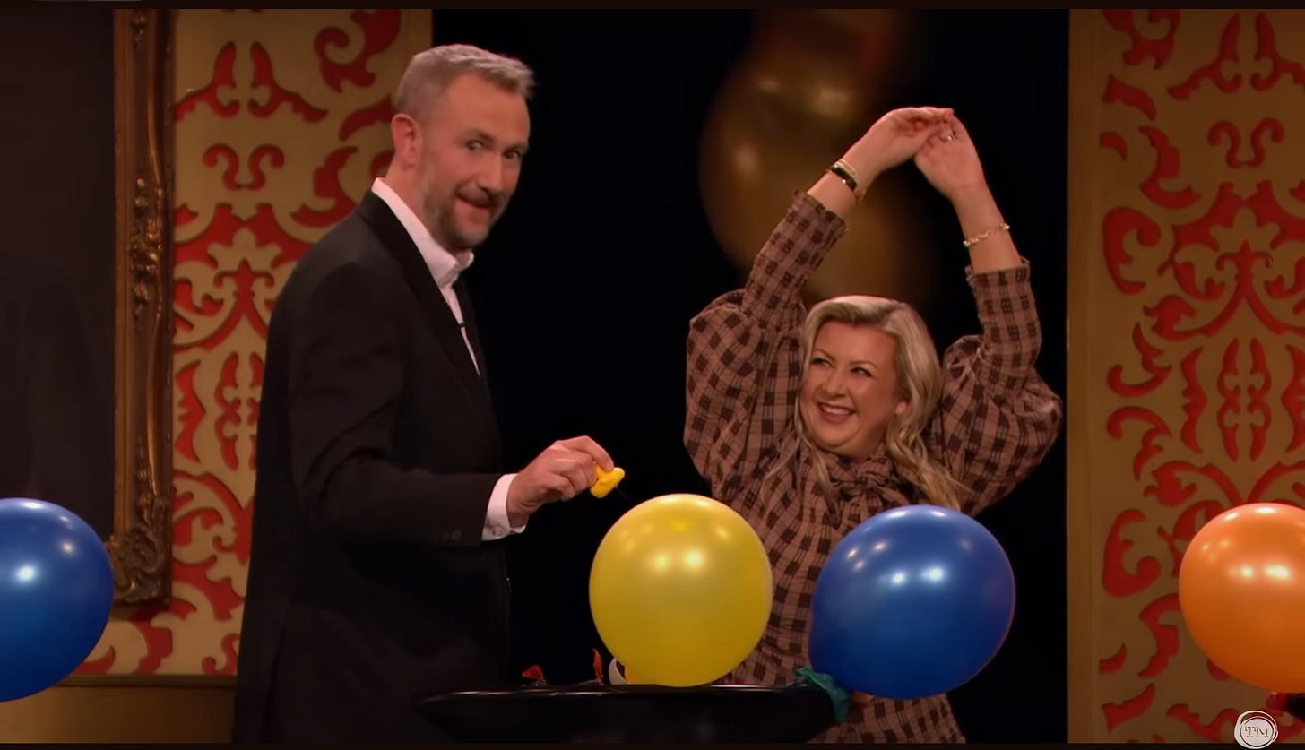 Rosie Ramsey dances with her arms up, looking alluringly at Little Alex Horne, who is about to pop colorful balloons.