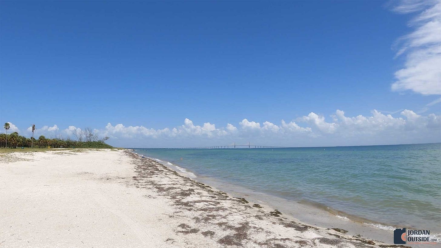 A wide view of East Beach at Fort De Soto Park, showing pale sand, scattered seaweed, and calm turquoise water stretching toward the Sunshine Skyway Bridge in the distance under a bright blue sky.