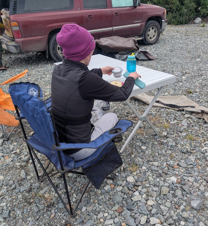 A girl in a bright magenta beanie and black jacket sits in a blue folding camp chair at a small white camp table, spreading something from a plastic container onto a bagel. A blue water bottle and a phone in a teal case sit on the table. Behind her, a maroon SUV is parked on the gravel with camp gear piled nearby. The ground is loose grey river rock in flat, overcast light.