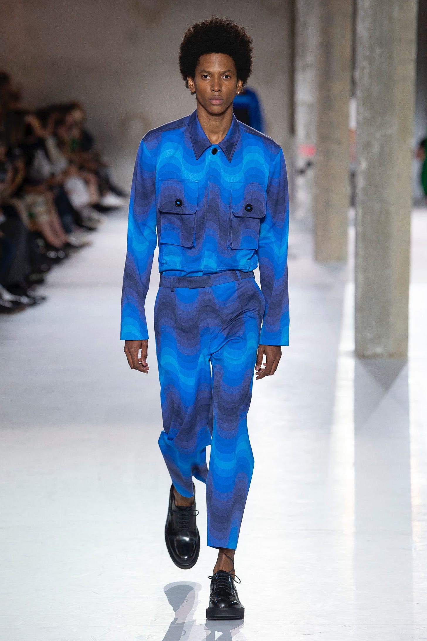 A model on a fashion catwalk, who is a Black man with a textured Afro, wearing a matching jackets and trousers characterised by a vibrant wavy graphic print in differing shades of blue, and black Derby shoes with a chunky sole. A model on a fashion catwalk, who is a Black man with a textured Afro, wearing a matching jackets and trousers characterised by a vibrant wavy graphic print in differing shades of blue, and black Derby shoes with a chunky sole.