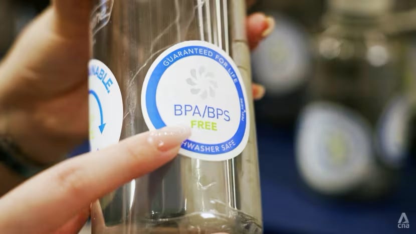 Close-up of a hand holding a glass bottle with a label reading "BPA/BPS Free," "Guaranteed for Life," and "Dishwasher Safe" in a store setting.