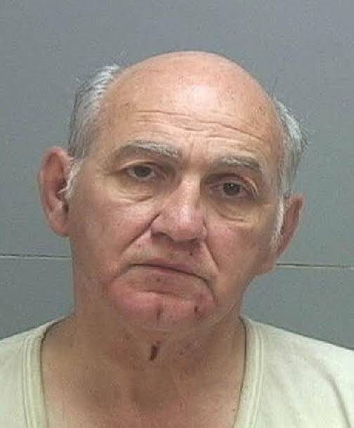 George Zinn, an elderly man with a bald head and gray hair on the sides, wearing a light-colored undershirt. He stands against a plain gray background.