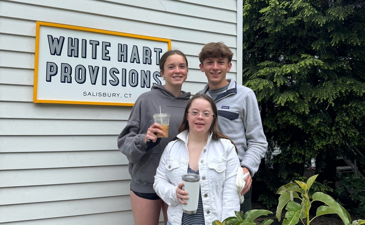 Marilee, Penny, and William stand outside a building with white siding next to a sign that reads “White Hart Provisions, Salisbury, CT.” Marilee, Penny, and William stand outside a building with white siding next to a sign that reads “White Hart Provisions, Salisbury, CT.”