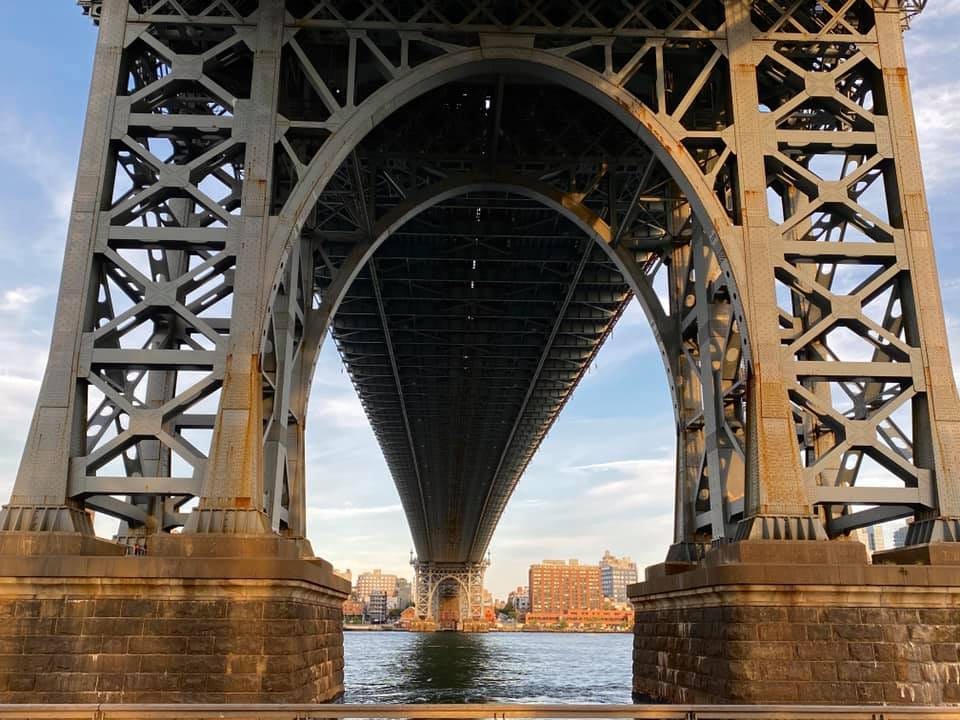 Looking up at the metal underside of the Manhattan Bridge from East River Park. There is blue sky and thin clouds above and the East River below. Looking up at the metal underside of the Manhattan Bridge from East River Park. There is blue sky and thin clouds above and the East River below.