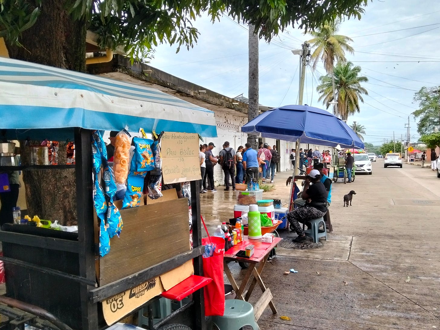 People stand in line on a sidewalk by a vendor cart and tables