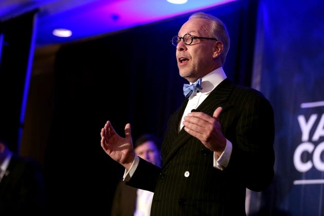 Jeffrey Tucker in a pinstripe suit and light blue bow tie speaks on stage under blue lighting. Jeffrey Tucker in a pinstripe suit and light blue bow tie speaks on stage under blue lighting.
