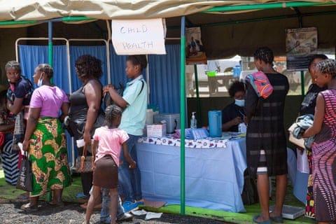 people stand in a line in front of a temporary structure bearing a hand written sign that reads ‘child health’.