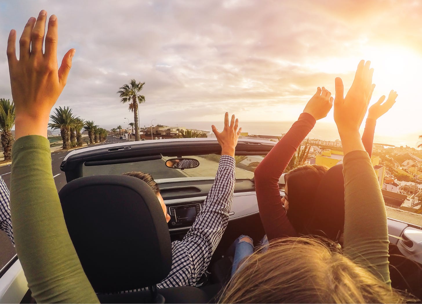 Rear view of three people riding in a convertible with their hands raised in the air, driving into a colorful California sunset on a clear evening.