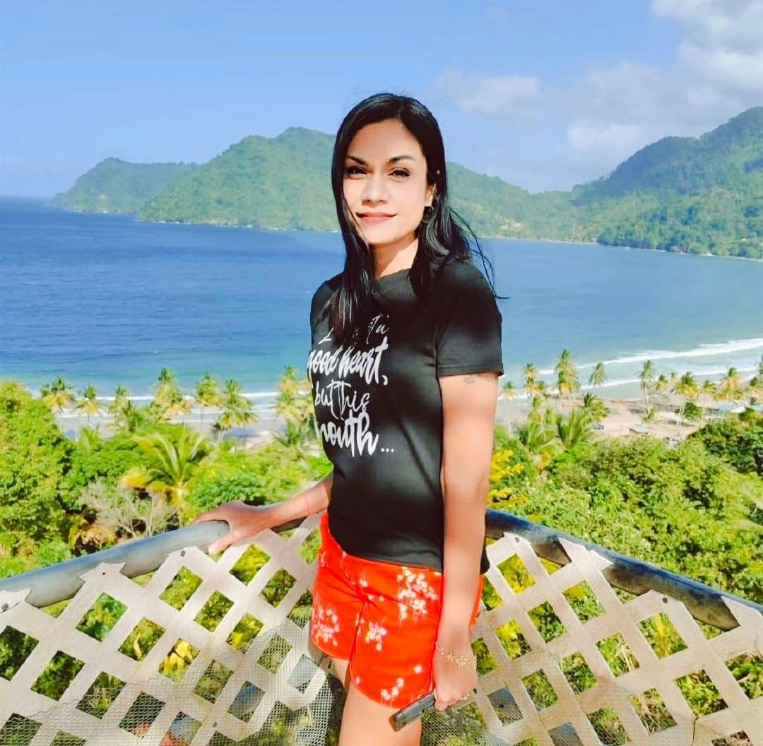 A person standing on a viewing platform overlooking a beautiful tropical coastline. They're wearing a black t-shirt with white text and bright orange shorts, with lush green mountains and a blue ocean bay in the background. Palm trees line the beach below, and the scene has clear blue skies with some clouds over the distant mountains.