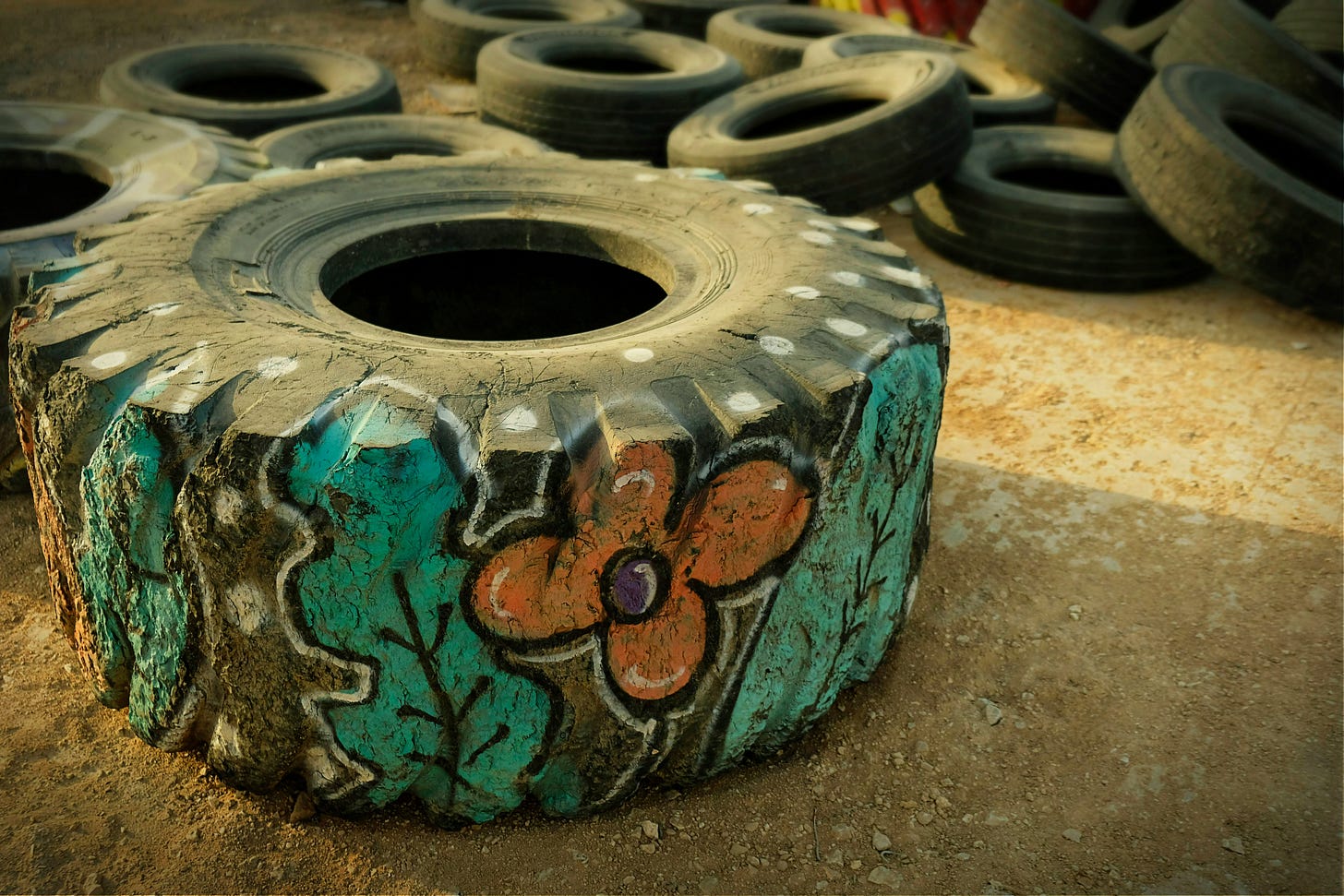 A wide black tractor tire on the dirt ground, painted with green leaves and orange flowers. There is a stack of other tires in the background.