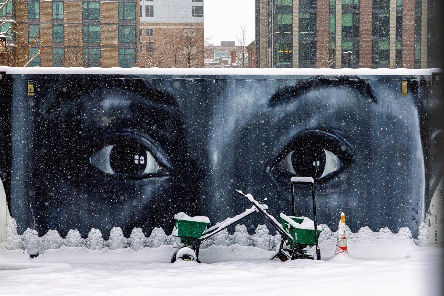 Snow-covered mural of human eyes on a construction barrier during a winter storm in Boston Seaport.