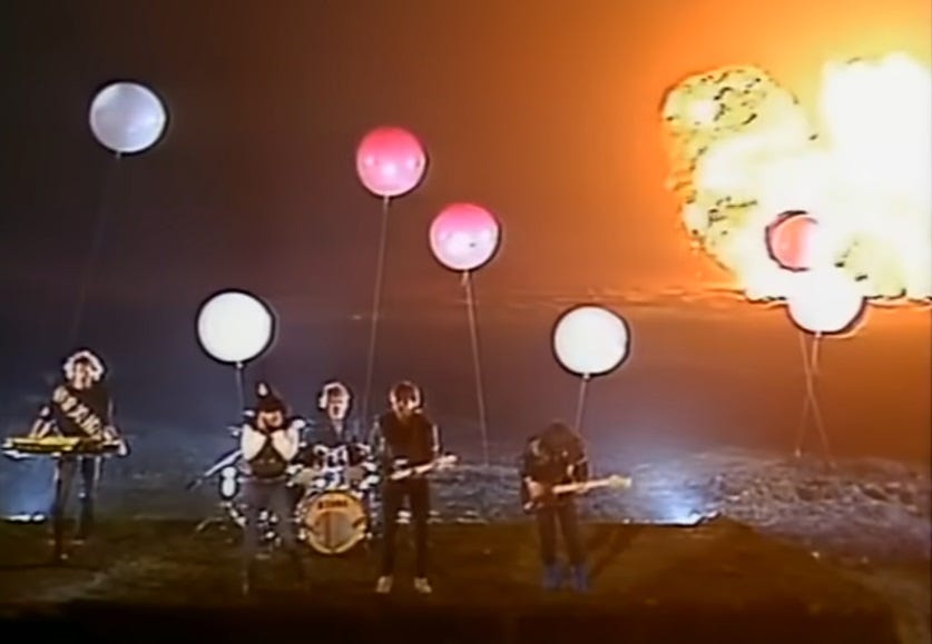 screenshot from Nena's '99 Luftballons' video (1983), showing the singer and her band on a stage in a desert landscape with multicolored balloons, as pyrotechnic explosions go off in the background. 