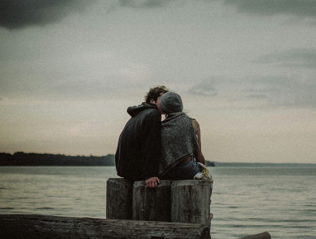 man and woman sitting and kissing on post near the body of water