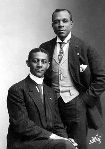 a black and white photo of two people in suits. One is sitting while the other is standing with his hands in his pockets