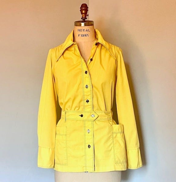 May include: A yellow long-sleeved button-up shirt with a belt and pockets. The shirt has a collar and is on a mannequin. The mannequin has the words 'Royal Form' printed on the chest.