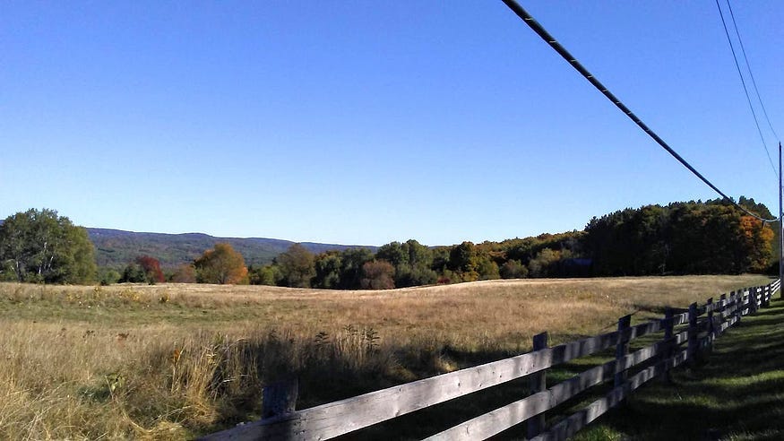 Hazy hills along the distant horizon, with a dry grassy field in the middle ground, while a plank fense and telegraph wire reach across the foreground.