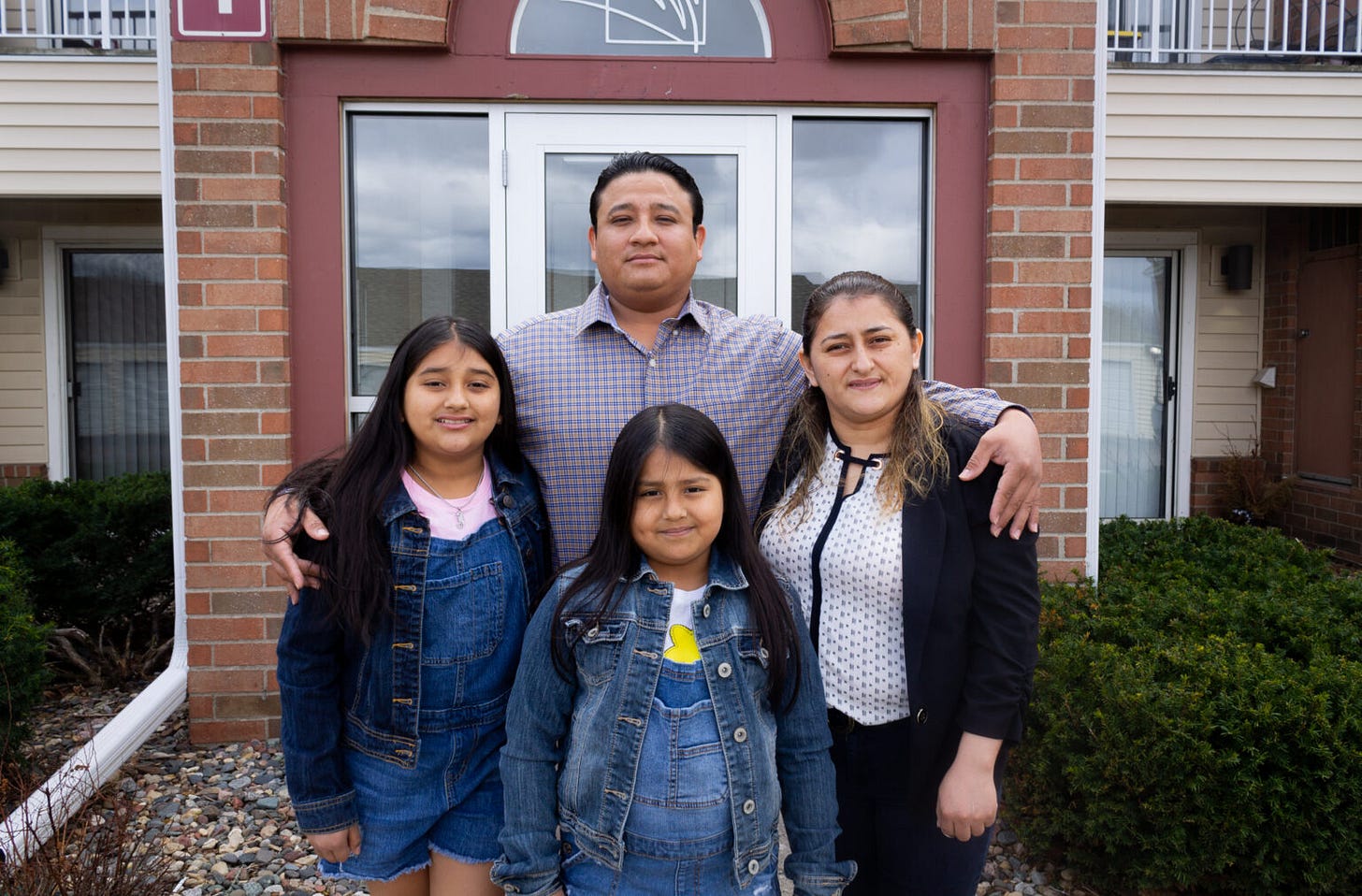 a family, two parents and two daughters wearing denim jackets stands in front of the front door of a residential building