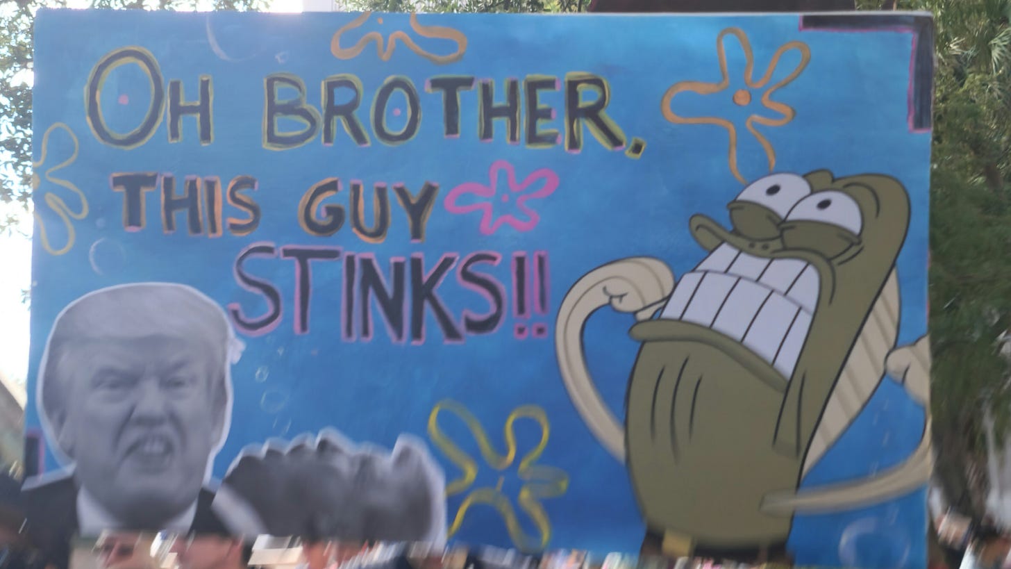 A blue hand-painted sign with a picture of Donald Trump and a SpongeBob character, reading "OH BROTHER, THIS GUY STINKS!!"