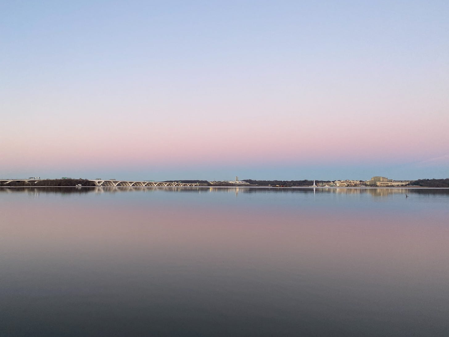 Photo of a calm, flat, wide river with a bridge in the distance and the sunset hues of blue, white, pink and purple reflected onto the water from the sky. Photo of a calm, flat, wide river with a bridge in the distance and the sunset hues of blue, white, pink and purple reflected onto the water from the sky.