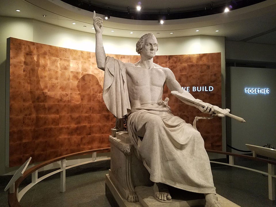 Smithsonian color photo of sculpture depicting a stern-faced, bare-chested, muscular George Washington in Roman attire, seated on an ornate chair. Right arm is raised with one finger pointing, as if giving a decree, while the left had holds out a sheathed sword, handle first. Smithsonian color photo of sculpture depicting a stern-faced, bare-chested, muscular George Washington in Roman attire, seated on an ornate chair. Right arm is raised with one finger pointing, as if giving a decree, while the left had holds out a sheathed sword, handle first.