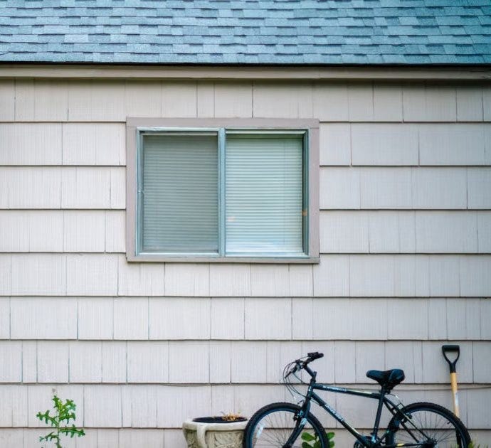 The side of a small house with off-white shingle siding, a window with closed blinds and a plain gray-blue shingle roof, with a bicycle and a shovel leaning against the wall beside a concrete planter