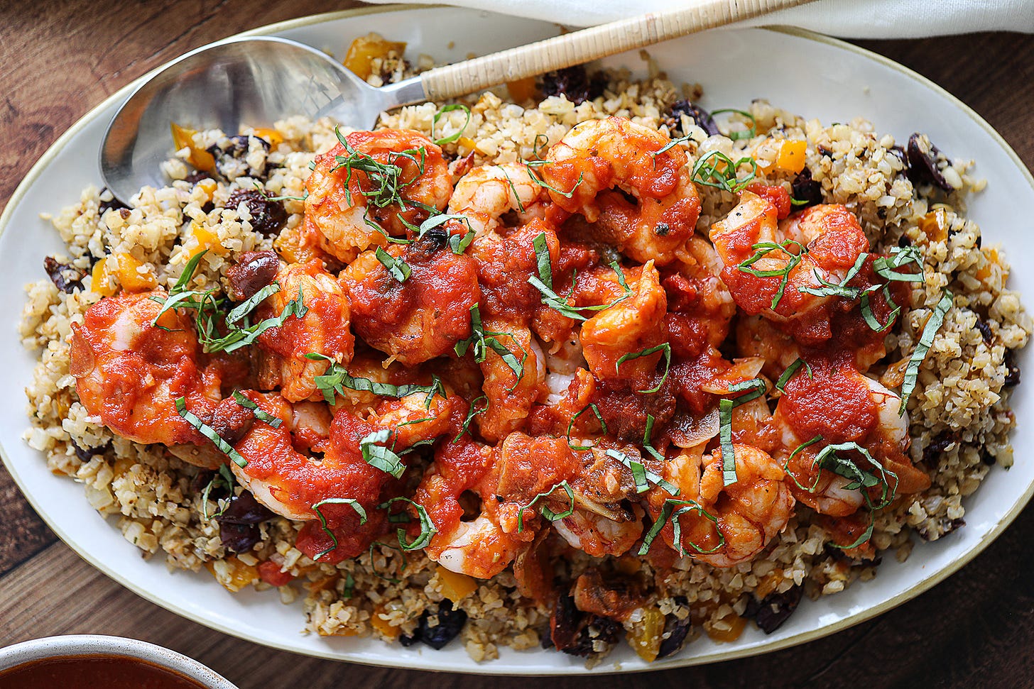 an oval white platter with Shrimp Puttanesca with Mediterranean Cauliflower Rice, the cauliflower rice is dotted with black Kalamata olives and the shrimp is covered with red puttanesca sauce and garnished with ribbons of fresh basil, and there's a stainless steel serving spoon with a bamboo handle wedged into the platter contents